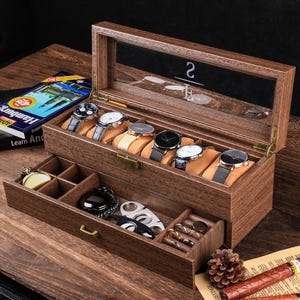 May include: A wooden watch box with a glass top, displaying several wristwatches on tan cushions. The box has a drawer with compartments for other accessories. The watches have black, silver, and gold tones. The box is on a wooden surface.