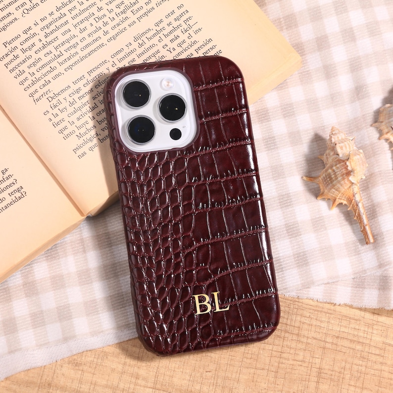 May include: A burgundy phone case with a crocodile skin texture. The case has a camera cutout and the initials "BL" in gold. The phone case is on a light-colored surface with a book and seashells.