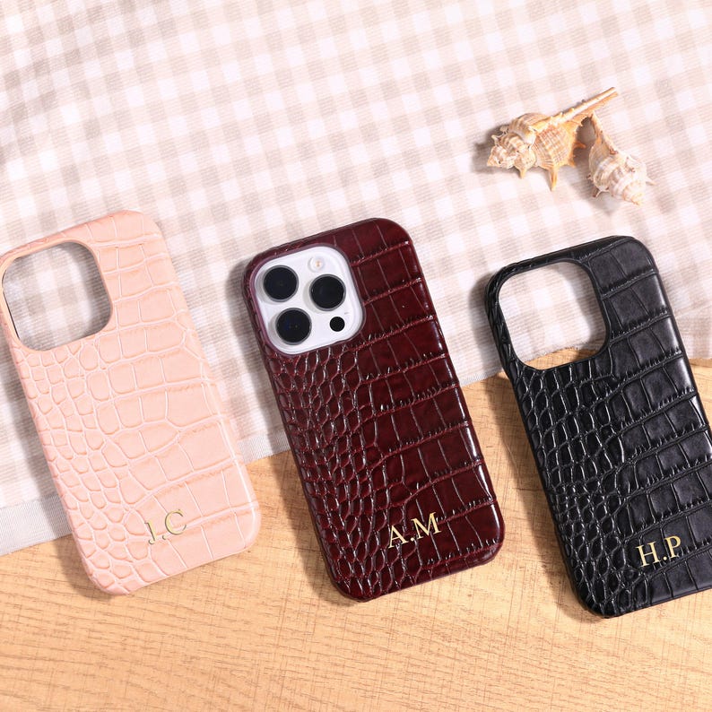 May include: Three personalized phone cases in pink, burgundy, and black, each with a crocodile skin texture. The cases have cutouts for the camera and are engraved with gold initials: J.C., A.M., and H.P. The cases are displayed on a light-colored surface.