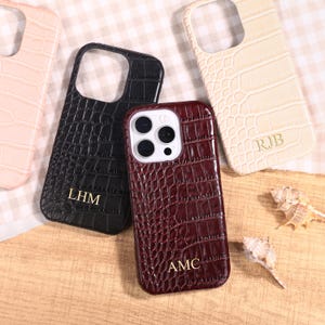 May include: A collection of personalized phone cases in various colors, including burgundy, black, and cream. Each case features a textured, crocodile-like pattern and initials. Decorative seashells are scattered around the cases.