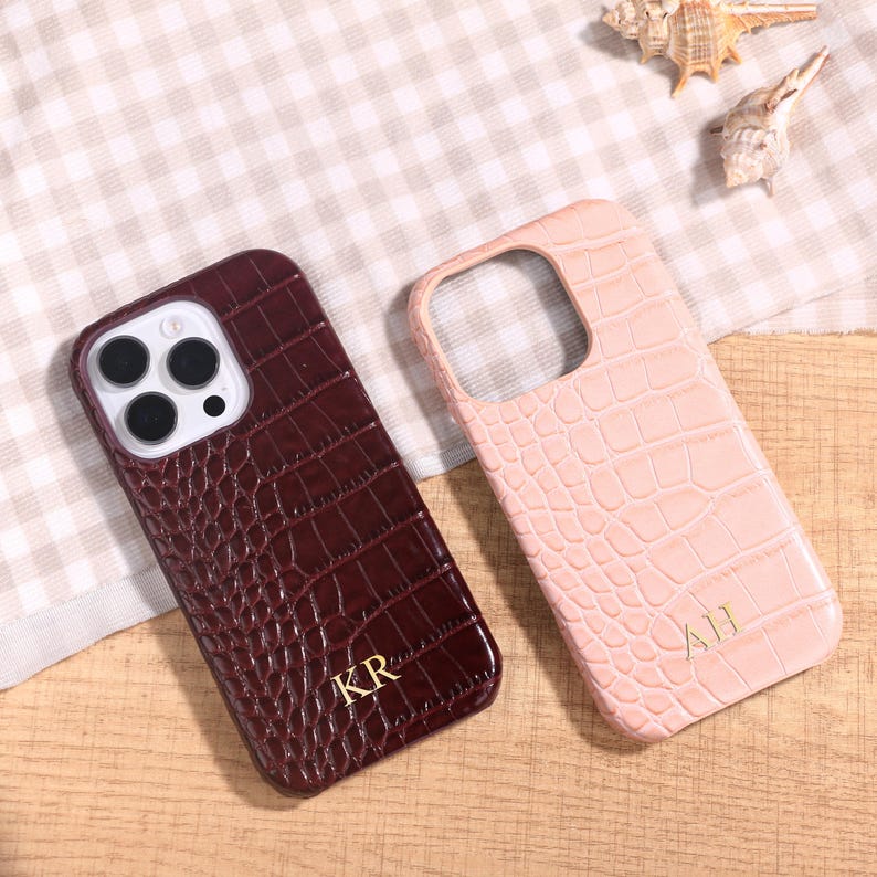 May include: Two phone cases with a crocodile skin texture. One is a deep burgundy color with the initials "KR" in gold. The other is a light pink with the initials "AH" in gold. Both cases are on a wooden surface.