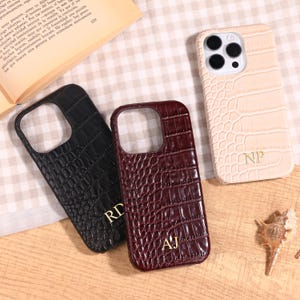 May include: Three phone cases with a crocodile skin texture. One is black, one is burgundy, and one is beige. Each case has a different set of initials in gold. The cases are on a light-colored, patterned surface.