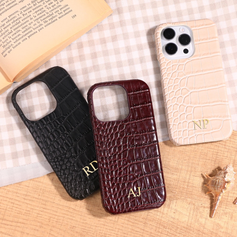 May include: Three phone cases with a crocodile skin texture. One is black, one is burgundy, and one is beige. Each case has a different set of initials in gold. The cases are on a light-colored, patterned surface.