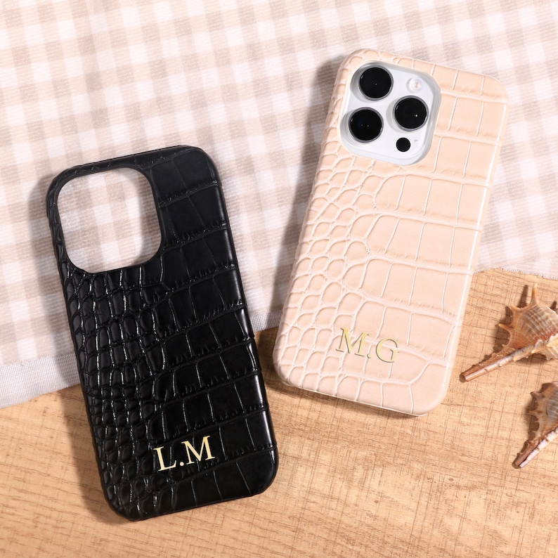 May include: Two phone cases with a crocodile skin texture. One is black with gold lettering "L.M", the other is beige with gold lettering "M.G". The cases are on a wooden surface with a checkered background.