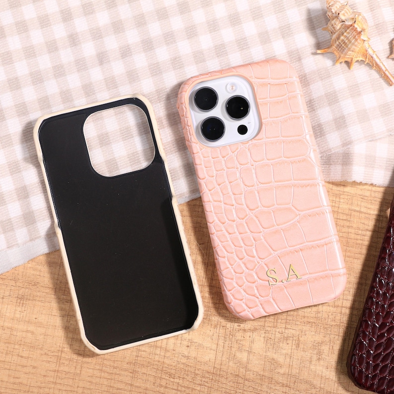 May include: A light pink phone case with a crocodile skin texture and the initials "S.A." in gold. The case is next to a black phone case with a white border. A seashell is in the background.
