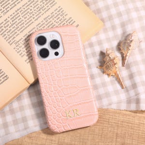 May include: A light pink phone case with a crocodile skin texture, featuring the initials "KR" at the bottom. The phone case is next to an open book and two seashells.
