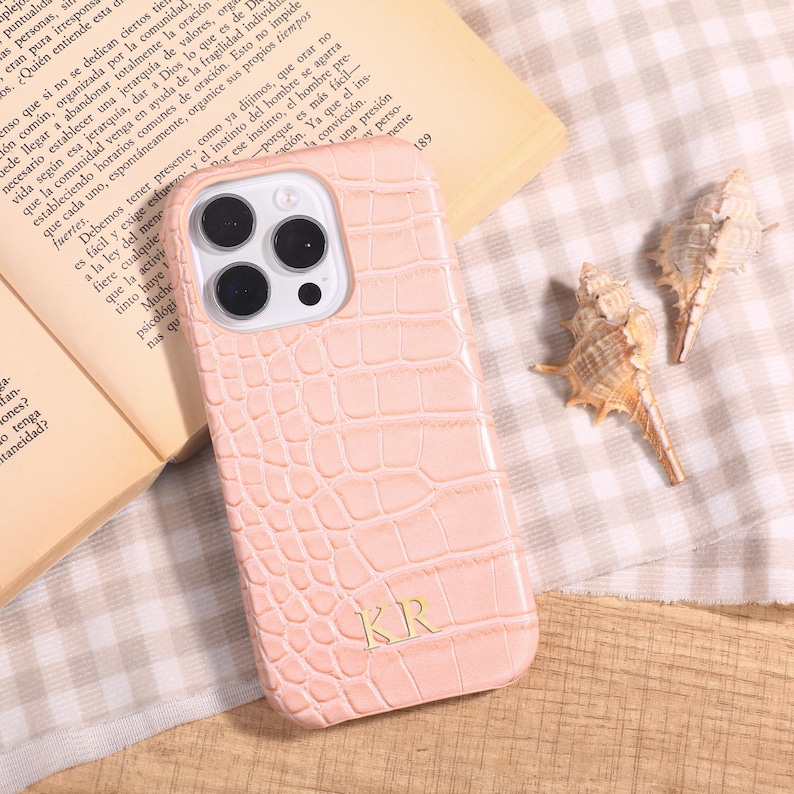 May include: A light pink phone case with a crocodile skin texture, featuring the initials "KR" at the bottom. The phone case is next to an open book and two seashells.