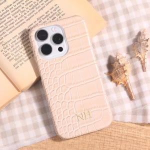 May include: A light beige phone case with a crocodile skin texture. The case has a camera cutout and the initials "NH" in gold. The case is lying on a patterned surface with a book and seashells.
