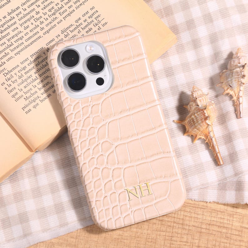 May include: A light beige phone case with a crocodile skin texture. The case has a camera cutout and the initials "NH" in gold. The case is lying on a patterned surface with a book and seashells.