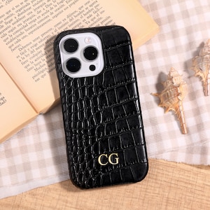 May include: Black phone case with a crocodile skin texture. The case has a gold-colored monogram of the letters "CG" on the lower right. The phone case is shown on a patterned surface with a book and seashells in the background.