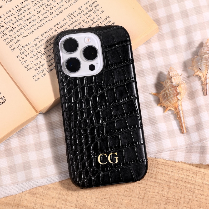 May include: Black phone case with a crocodile skin texture. The case has a gold-colored monogram of the letters "CG" on the lower right. The phone case is shown on a patterned surface with a book and seashells in the background.