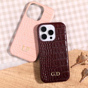 May include: Two phone cases with a crocodile skin texture. One is light pink with gold initials "LP", the other is burgundy with gold initials "G.D". The cases are on a wooden surface.