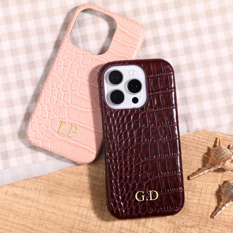 May include: Two phone cases with a crocodile skin texture. One is light pink with gold initials "LP", the other is burgundy with gold initials "G.D". The cases are on a wooden surface.