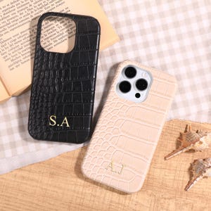 May include: Two phone cases with a crocodile skin texture are displayed. One is black with gold initials "S.A", the other is beige with gold initials "A.J". The cases are designed to protect the phone.