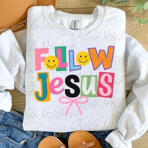 May include: A light gray sweatshirt with the words "FOLLOW JESUS" in colorful, block letters. The letters are in various colors, including pink, orange, green, and blue, with smiley faces and a pink bow.