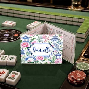 May include: A personalized Mahjong card holder with a floral and pagoda design. The holder features the name "Danielle" in a blue frame. The background includes pink flowers, blue pagodas, and a bridge, set against a white backdrop.