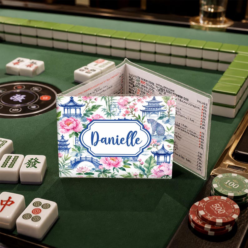 May include: A personalized Mahjong card holder with a floral and pagoda design. The holder features the name "Danielle" in a blue frame. The background includes pink flowers, blue pagodas, and a bridge, set against a white backdrop.