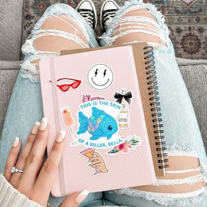 May include: A pink notebook adorned with stickers, including a smiling face, red sunglasses, and a blue fish. The text "THIS IS THE SKIN OF A KILLER, BELLA" is visible. A brown spiral-bound notebook rests beside it.