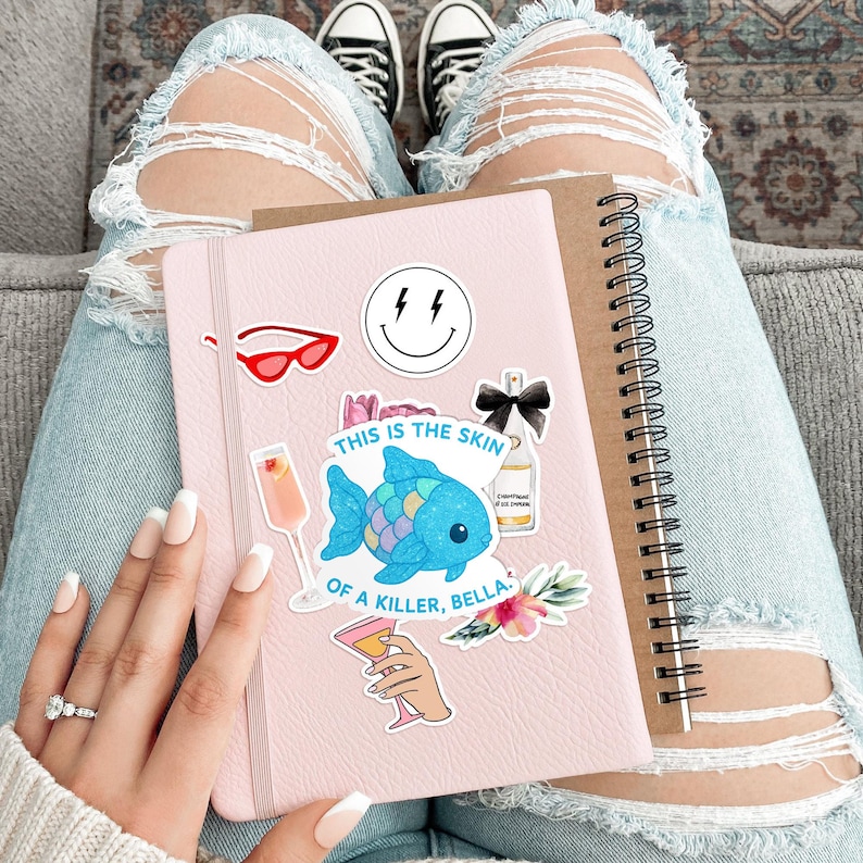 May include: A pink notebook adorned with stickers, including a smiling face, red sunglasses, and a blue fish. The text "THIS IS THE SKIN OF A KILLER, BELLA" is visible. A brown spiral-bound notebook rests beside it.