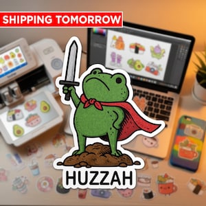 May include: A green frog sticker with a red cape and a sword, standing on a mound of dirt. The sticker has the word "Huzzah" at the bottom. The text "SHIPPING TOMORROW" is at the top.