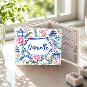 May include: A personalized, clear acrylic menu holder featuring a floral and pagoda design with the name "Danielle" in a blue frame. The design includes pink flowers, blue pagodas, and a bridge.