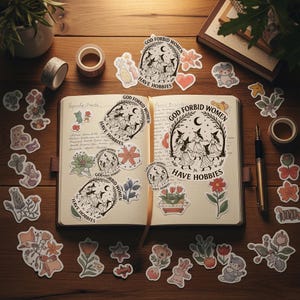 Puede incluir: Un diario abierto con pegatinas y cinta adhesiva decorativa sobre una superficie de madera. Las pegatinas presentan ilustraciones de flores, animales y el texto "GOD FORBID WOMEN HAVE HOBBIES". Tambi&eacute;n se ven un bol&iacute;grafo y una peque&ntilde;a planta en maceta.