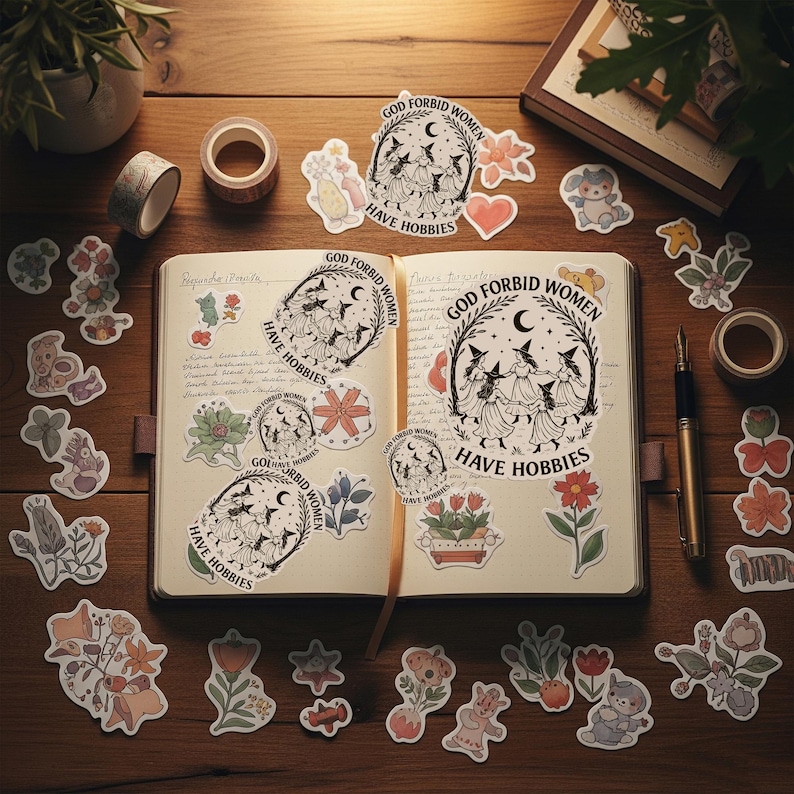 Puede incluir: Un diario abierto con pegatinas y cinta adhesiva decorativa sobre una superficie de madera. Las pegatinas presentan ilustraciones de flores, animales y el texto "GOD FORBID WOMEN HAVE HOBBIES". Tambi&eacute;n se ven un bol&iacute;grafo y una peque&ntilde;a planta en maceta.