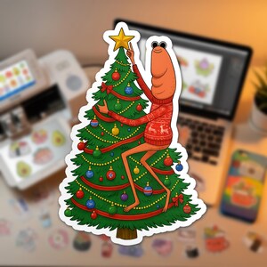 May include: A sticker featuring a cartoon shrimp wearing a red sweater with reindeer designs, placing a star on top of a decorated Christmas tree. The tree is adorned with ornaments and red ribbon.