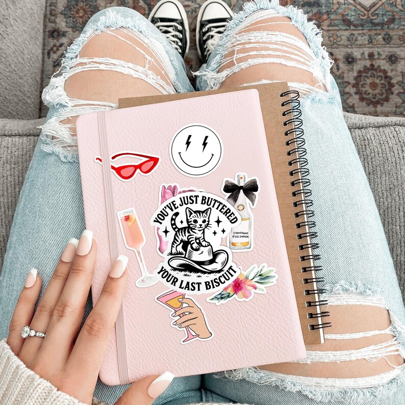May include: A pink notebook adorned with stickers, including a smiling face, red sunglasses, and a cat with the text "You've Just Buttered Your Last Biscuit." A brown spiral-bound notebook rests beside it. The image is set against a background of ripped blue jeans.