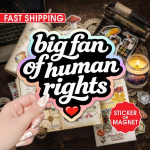 May include: A sticker or magnet with the text "big fan of human rights" in a black, bubble-style font, outlined with a rainbow gradient. A red heart is at the bottom. The background includes a candle, stickers, and a laptop.