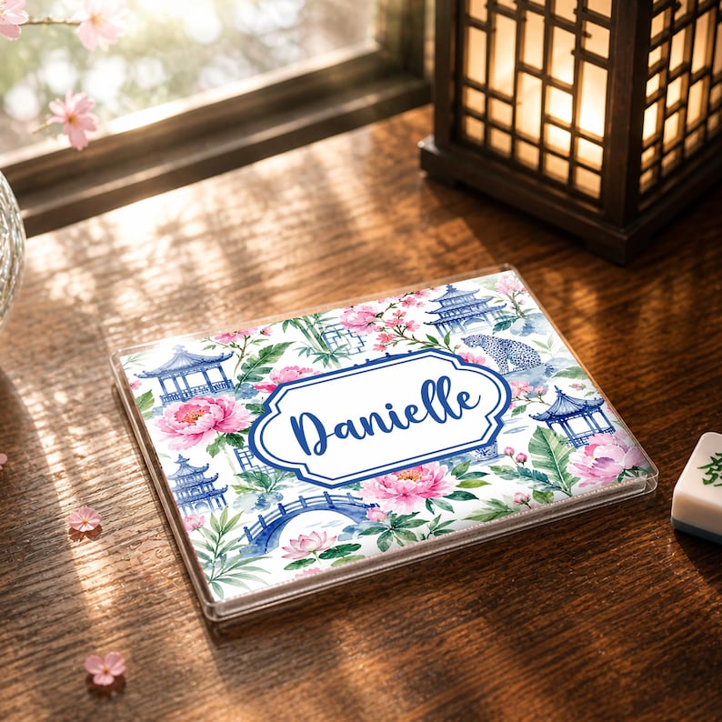 May include: A clear, rectangular case with a floral design featuring pink peonies, blue pagodas, and a bridge. The name "Danielle" is written in blue script within a white and blue frame. The case is on a wooden surface.