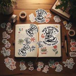 May include: A collection of stickers, washi tape, and a journal on a wooden surface. The stickers feature various floral and animal designs, along with a cat illustration and the text "YOU'VE JUST BUTTERED YOUR LAST BISCUIT".