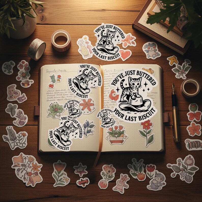 May include: A collection of stickers, washi tape, and a journal on a wooden surface. The stickers feature various floral and animal designs, along with a cat illustration and the text "YOU'VE JUST BUTTERED YOUR LAST BISCUIT".