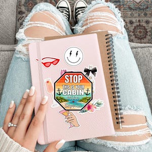 May include: A pink notebook with various stickers, including a smiley face, sunglasses, and a sign that reads "STOP THIS IS YOUR CABIN". A brown spiral notebook is next to it. The person is wearing ripped blue jeans and black and white sneakers.