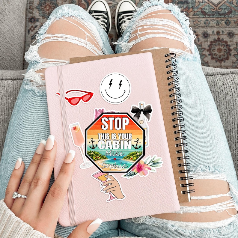 May include: A pink notebook with various stickers, including a smiley face, sunglasses, and a sign that reads "STOP THIS IS YOUR CABIN". A brown spiral notebook is next to it. The person is wearing ripped blue jeans and black and white sneakers.