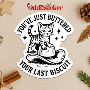 May include: A white sticker with a black and white illustration of a cat standing on a cowboy hat. The sticker has the text "YOU'VE JUST BUTTERED YOUR LAST BISCUIT" in a circular design. The sticker has a decorative border.