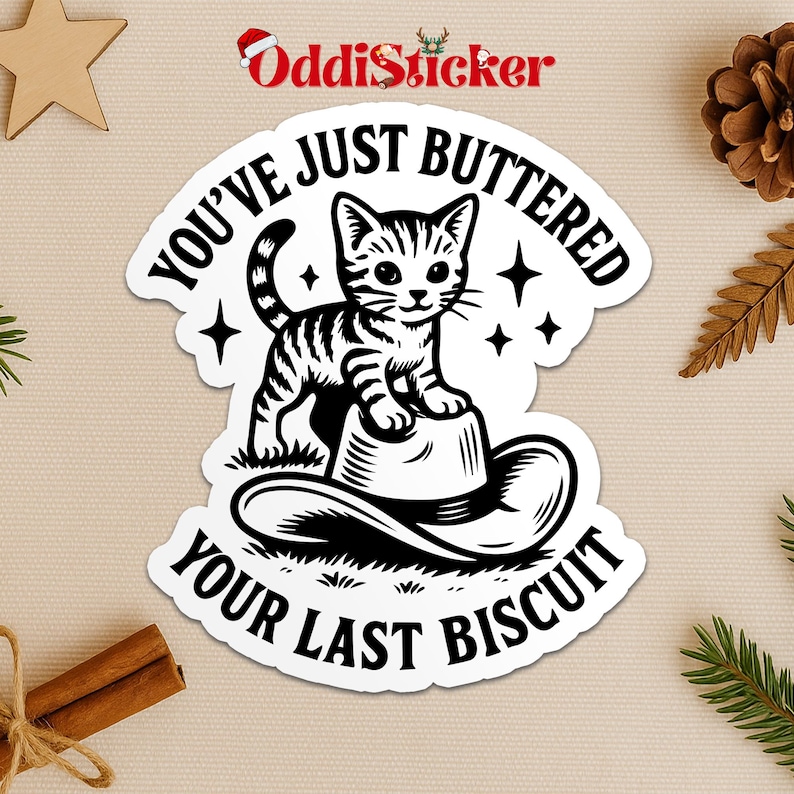 May include: A white sticker with a black and white illustration of a cat standing on a cowboy hat. The sticker has the text "YOU'VE JUST BUTTERED YOUR LAST BISCUIT" in a circular design. The sticker has a decorative border.