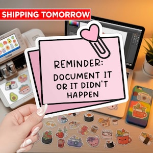 May include: A pink sticky note with the text "REMINDER: DOCUMENT IT OR IT DIDN'T HAPPEN" is held by a hand. A variety of colorful food-themed stickers are scattered on a wooden surface. A laptop and a Brother cutting machine are in the background.