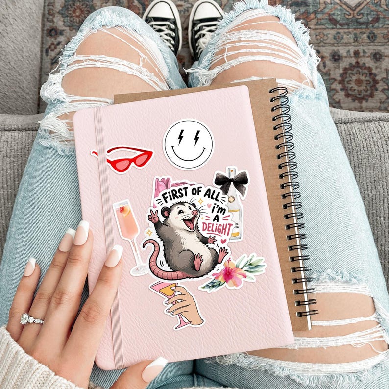 Puede incluir: Un cuaderno rosa adornado con pegatinas, incluyendo una zarig&uuml;eya sonriente, gafas de sol rojas y una carita sonriente. Un cuaderno marr&oacute;n con espiral est&aacute; al lado. La persona lleva vaqueros azules rotos y zapatillas negras.