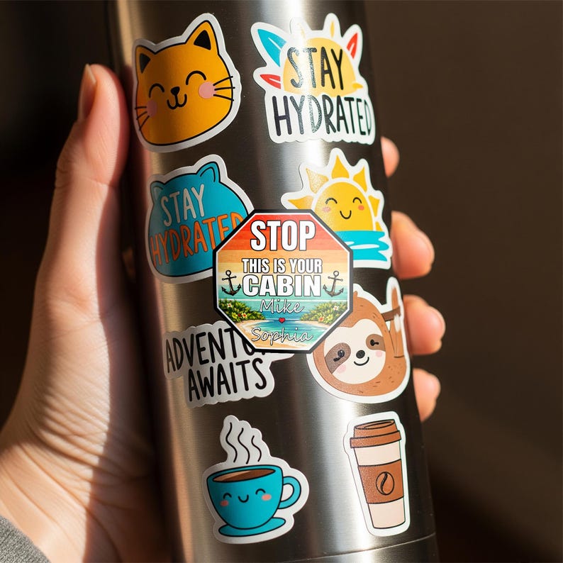 May include: A collection of colorful stickers on a stainless steel water bottle. The stickers feature various designs, including a smiling cat, a sun, a sloth, a coffee cup, and the text "Stay Hydrated" and "Adventure Awaits".