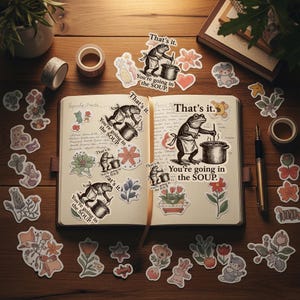 May include: An open notebook with handwritten notes and decorative stickers scattered around. The stickers feature a frog chef with the text "You're going in the SOUP" and "That's it." Floral and animal-themed stickers are also present.