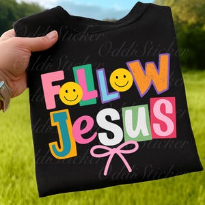 May include: Black crew neck t-shirt with the words "FOLLOW JESUS" in colorful, block letters. The design includes smiley faces and a pink bow. The shirt is held up against a green background.