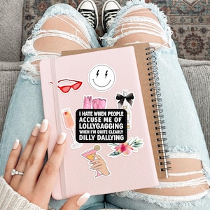 May include: A pink notebook with various stickers, including a smiley face, sunglasses, and cocktail illustrations. The notebook has the text "I hate when people accuse me of lollygagging when I'm quite clearly dilly dallying."