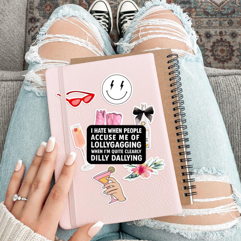 May include: A pink notebook with various stickers, including a smiley face, sunglasses, and cocktail illustrations. The notebook has the text "I hate when people accuse me of lollygagging when I'm quite clearly dilly dallying."