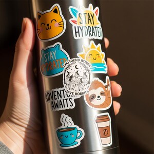 Puede incluir: Una botella de agua de acero inoxidable adornada con una variedad de pegatinas coloridas. Las pegatinas presentan un gato sonriente, un sol, un perezoso, una taza de caf&eacute; y los textos "Stay Hydrated", "Adventure Awaits" y "God Forbid Women Have Hobbies".