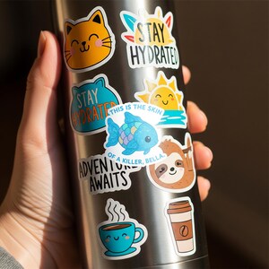 May include: A collection of colorful vinyl stickers with various designs, including a smiling cat, a sun, a sloth, a fish, and coffee cups. Text includes "Stay Hydrated," "Adventure Awaits," and "This is the skin of a killer, Bella."