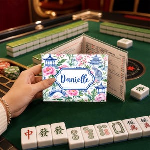 May include: A personalized Mahjong card holder with a floral and pagoda design. The card holder features the name "Danielle" in a blue frame. Pink flowers and blue pagodas are set against a white background. The holder is open, revealing a price list.