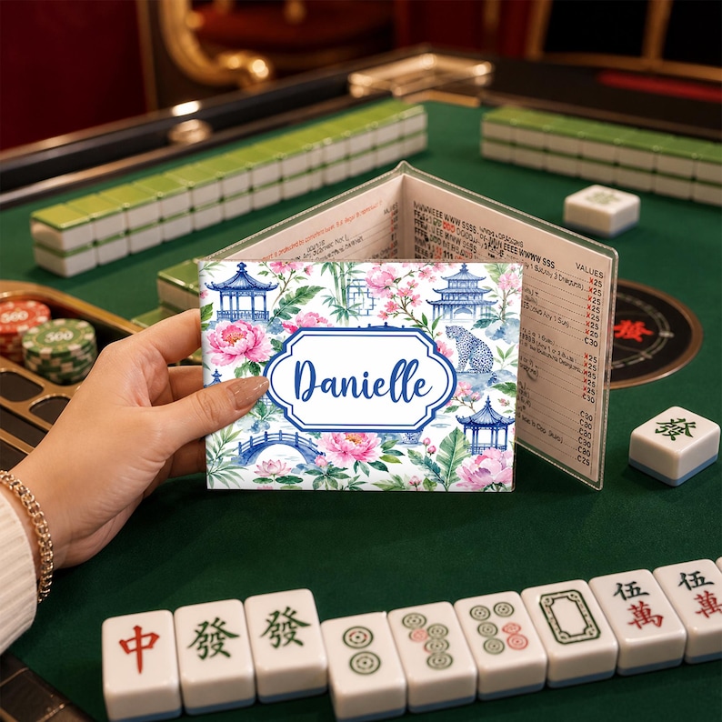 May include: A personalized Mahjong card holder with a floral and pagoda design. The card holder features the name "Danielle" in a blue frame. Pink flowers and blue pagodas are set against a white background. The holder is open, revealing a price list.