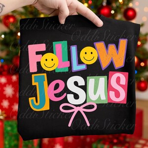 May include: Black sweatshirt with the words "FOLLOW JESUS" in colorful, block letters. The design includes smiley faces and a pink bow. The sweatshirt is held up by a hand.
