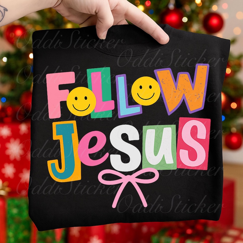 May include: Black sweatshirt with the words "FOLLOW JESUS" in colorful, block letters. The design includes smiley faces and a pink bow. The sweatshirt is held up by a hand.